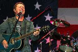 A dark-haired man playing a guitar and singing into a microphone in front of the American flag