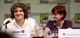 A Caucasian male wearing a white shirt sits in front of a microphone, smiling. He has large, curly brown hair which almost covers his eyes. Beside him is a Caucasian woman with short, dyed-red hair, smiling as well and looking at the man next to her.