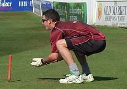 A man dressed in a dark red training top and black shorts with a dark red stripe crouches behind a stump with his gloved hands poised in front of him.