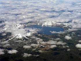 The caldera of Mount Mazama is visible, filled with the water of Crater Lake. A mountain sits to the left of the lake. Snow surrounds the lake and sits atop the nearby mountain.