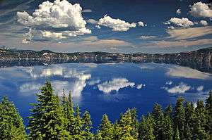 The water of Crater Lake can be seen above a forested area in the foreground.