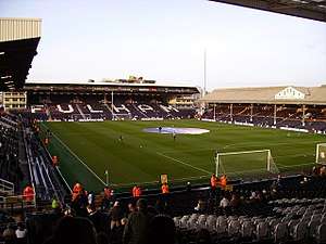 A colour photograph of Craven Cottage football stadium, showing the pitch in the centre of the image, surrounded stands. At the top of the photo, the word 'Fulham' is spelt out in white lettering on the black seats.