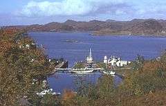 A small harbour surrounded by trees offers shelter to several yachts, fishing boats and a motor launch. The harbour is connected to the sea beyond by a lock gate. A small skerry lies in the water and brown rocky hills dominate the distance.