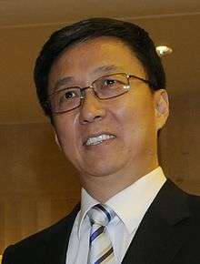 a smiling man with a short haircut, wearing glasses, a white shirt, a suit and a white tie with blue stripes