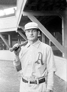 A man wearing an old-style white baseball uniform with an script "A" over the left breast and white pillbox cap holds a baseball bat over his right shoulder.