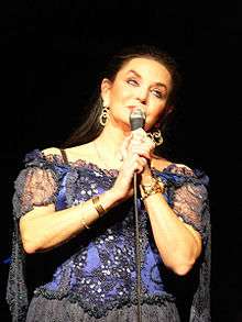 A woman with very long dark hair, wearing a purple and black lace dress, singing into a microphone
