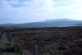 Photograph of a long, flat mountain ridge on the far horizon, with layers to the fore: foothills of the aforementioned mountain; a sliver of a lake; blanket bog and a fence in the very near foreground.