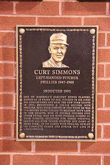 A bronze plaque mounted on a brick wall with the face of a smiling man wearing a baseball cap; the primary captions read "CURT SIMMONS; Left-handed pitcher; Phillies 1947&ndash;1960"