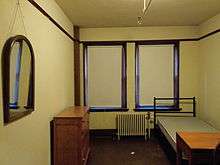 A room with dark floors, brown trim and, from left to right, a mirror hanging on the wall, a dresser, two windows with their shades drawn fully, a radiator, a bed, and a desk