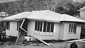 Black and white photograph of a contorted single-story home; a crushed automobile is visible beneath the structure