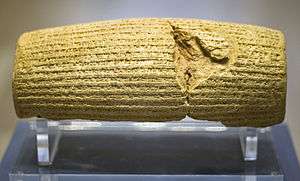 Front view of a barrel-shaped clay cylinder resting on a stand. The cylinder is covered with lines of cuneiform text