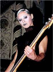 A worm's eye view shot of D'arcy Wretzky—a Caucasian female with short blond hair, wearing a black suit and black horns on her head—plays bass guitar on stage.