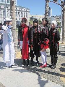 Five people doing cosplay of manga and anime series. While the first one is mostly dressed in white, the other four are dressed in black and red uniforms.