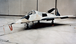 Small jet aircraft with angled surfaces in hangar. It is painted in a disruptive scheme to confuse casual onlookers.