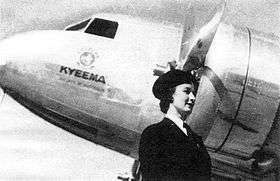 Half portrait of stewardess in dark cap and uniform in front of nose and engines of piston-engined airliner, emblazoned with the name Kyeema