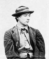A young man looking sour and facing right. He is dressed in a jacket, bow tie, and wide-brimmed hat, and his shirt is partially unbuttoned.