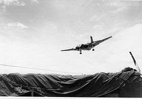 Large airplane descending over tarpaulin-covered area