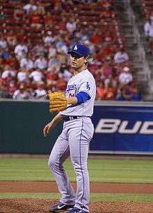 A man wearing a gray baseball uniform and blue baseball cap extends his baseball-gloved left hand.