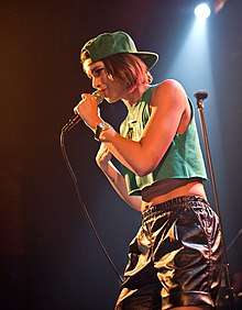 An image of a woman performing on a stage. She is wearing a crop top, shiny shorts, and a baseball cap. She is singing into a microphone and looking away from the camera.