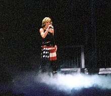 Faraway image of a woman in a short red skilt and black top standing on a stage. Smokes billow around her feet.