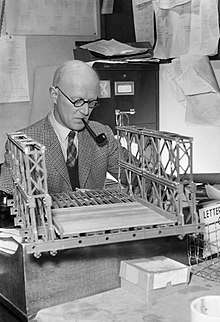 A middle-aged man sits at a desk while smoking a pipe; he is examining a model bridge in front of him