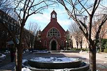 A frontal view of the red, brick edifice of the chapel. In front is a brick quadrangle with some snow on the ground, all centered between two trees in the foreground.