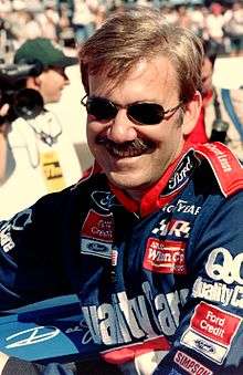A mustached man in his early forties wearing sunglasses and blue and red racing overalls