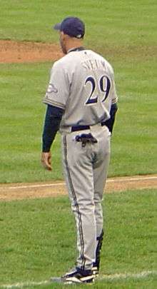 A man wearing a gray baseball uniform and navy-blue baseball cap facing away from the camera