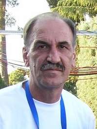 An elderly man with receding hairline and a moustache, wearing a white T-shirt with a blue ribbon down his neck.