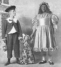 two men dressed as a boy and a girl hold the hands of a rag-doll between them