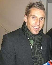 Smiling 30-ish white man with spiky hair wearing coat and scarf, pictured indoors near a door marked "Players Lounge". Two other men converse in the background.