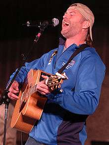 A man wearing a blue shirt and a reversed baseball cap, playing guitar and singing into a microphone