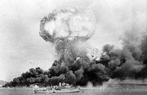 Long shot of mushroom cloud from an explosion, and black billowing smoke from nearby fire, with ship in foreground.