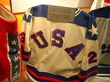 A white hockey jersey with the letters "U-S-A" on the front in blue with red trim. The jersey is inside a glass case.