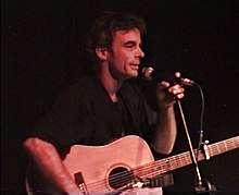 A man is shown in a right profile, upper body shot. He holds a microphone on a stand with his left hand close to his mouth. With his other hand he cradles an acoustic guitar. He wears a dark shirt and looks ahead.