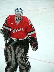 An ice hockey goaltender skates on ice. He is wearing a red and white sweater, and black and grey pads.
