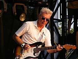 A closeup of Byrne playing guitar in a white suit and sunglasses