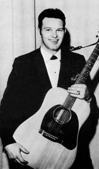 A dark-haired young man in a dark jacket, smiling broadly while holding a guitar
