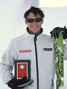 A man with brown hair, round head and wearing sunglasses is smiling broadly. He is wearing a white jacket and black gloves and is holding an award plaque in his right hand next to his midriff. His left hand is holding green-coloured skis.