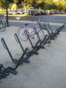 An array of sixteen or so parking spots for bicycles. Each is a square metal bar angled at 45 degrees from the ground with a parallel loop of thinner round tubing at the top. They are set in a row with the bars pointing in alternating directions. A pink ladies' bicycle with a shipping basket is parked in one. Another row of them is mostly out of frame to the left with a mountain bike parked in it. In the background is a car park with trees.