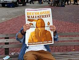 Occupier holding up newspaper, covering his face. Back of paper shows Native America, with caption "Decolonize WallStreeet, Decolonize the 99%"