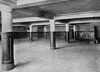 A black-and-white photo of an unfurnished basement, horizontally lit with diffuse light. It has a pale coffered ceiling, with thickish round pillars supporting the intersections of the beams. The lower half of both pillars and walls is covered with dark wood panelling. The bare floor is pale grey.