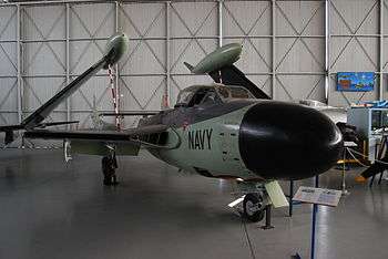 A green and black de Havilland Sea Venom jet-powered aircraft with wings folded up and "Navy" prominently printed on the fuselage