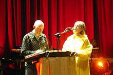 Upper body shot of a forty-five-year-old man at left. He is peering at a stringed instrument on a lectern. A microphone is directed to a forty-three-year-old woman at right. She is shown partly in left profile, singing with her eyes closed, her left hand rests on top of the lectern. She wears a ring on her left hand. In the background is musical equipment, a high stage curtain and a foot-light.