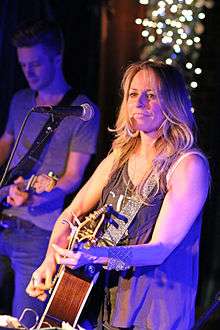 A woman with long blonde hair playing a guitar, with a man playing a guitar visible in the background
