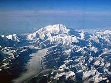 In an aerial image, a mountain is surrounded by many smaller mountains and a glacier