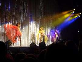 Three woman are shown to be performing; multi-colored lights shine upon them