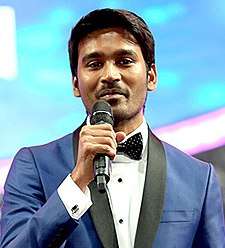 A picture of Dhanush with a Filmfare Award in his left hand as he looks at the camera and speaks.