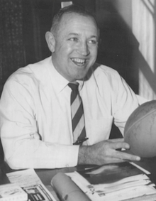 Richard F. Gallagher sitting at a table with a football in his hands in 1959
