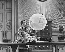 A black-and-white screenshot of a man sitting upon a table, holding a globe.